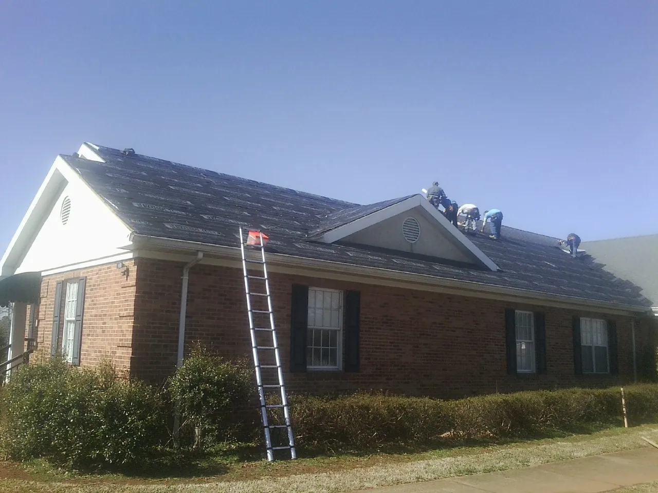 Crew tearing shingles off a brick ranch roof with a ladder set — Emergency Roof Repair work by our crew in Garden City