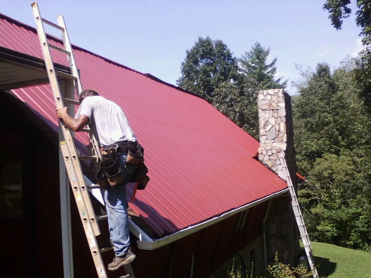 Roofer on a ladder beside a red metal roof and stone chimney — Soffit & Fascia Repair by Roofing Brothers in Garden City